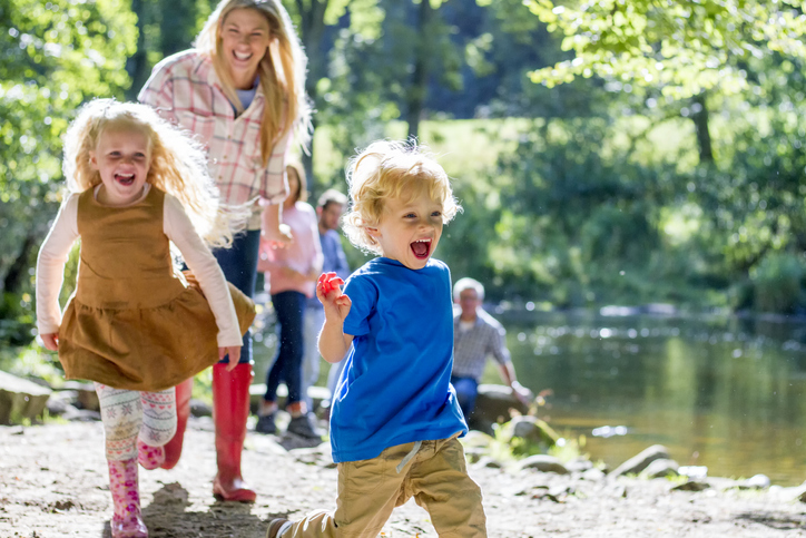 A family are sat at a lake on a nice summers day. The children are running ahead while parents look on.