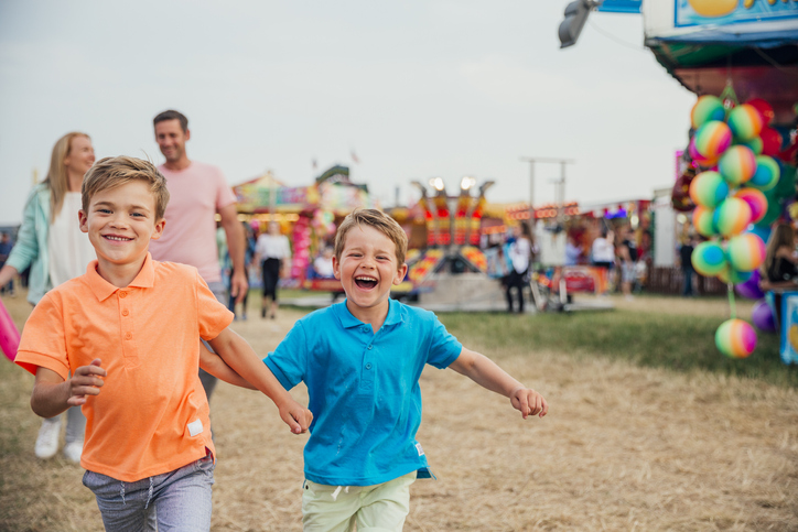 Family visit the fairground to enjoy the rides together. Little boys are running towards the camera. Parents walking behind.